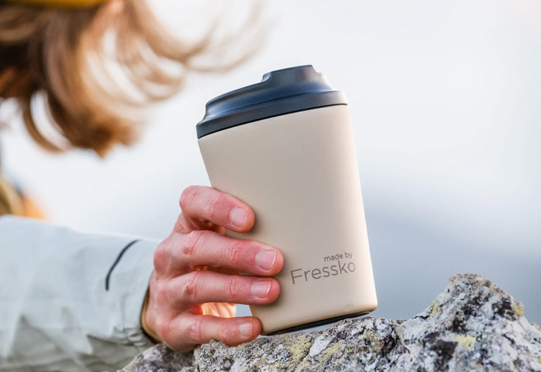 Person holding a Made By Fressko reusable coffee mug against a rock.
