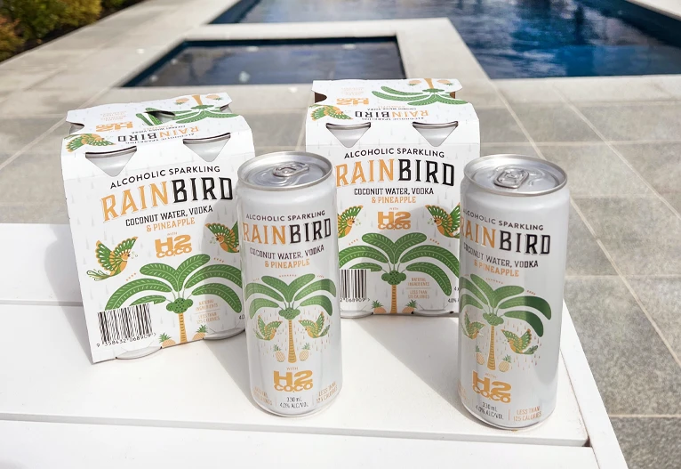 Rainbird H2Coco Vodka Pineapple premix products on a table next to a backyard pool.